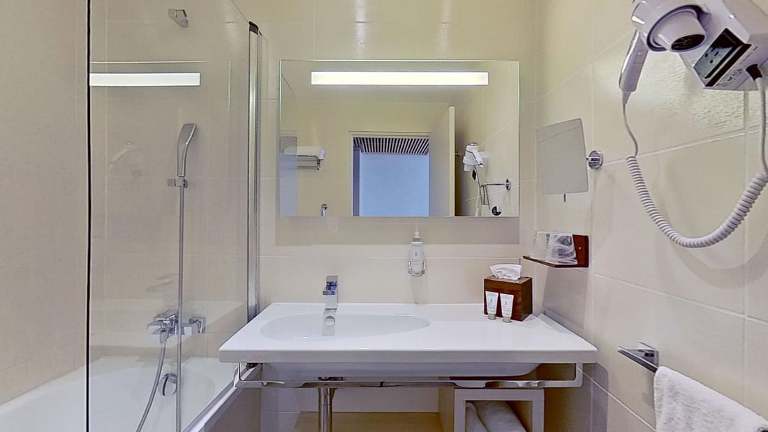 Modern bathroom with shower, sink, and mounted hairdryer, featuring a glass enclosure and white tiles.