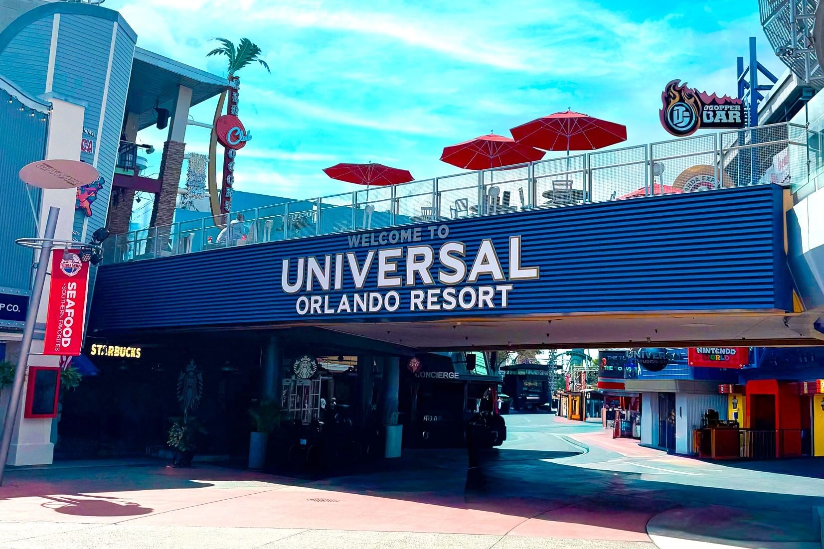 A walkway with a large sign that reads Universal Orlando Resort topped with patio chairs and red umbrellas at Universal CityWalk.