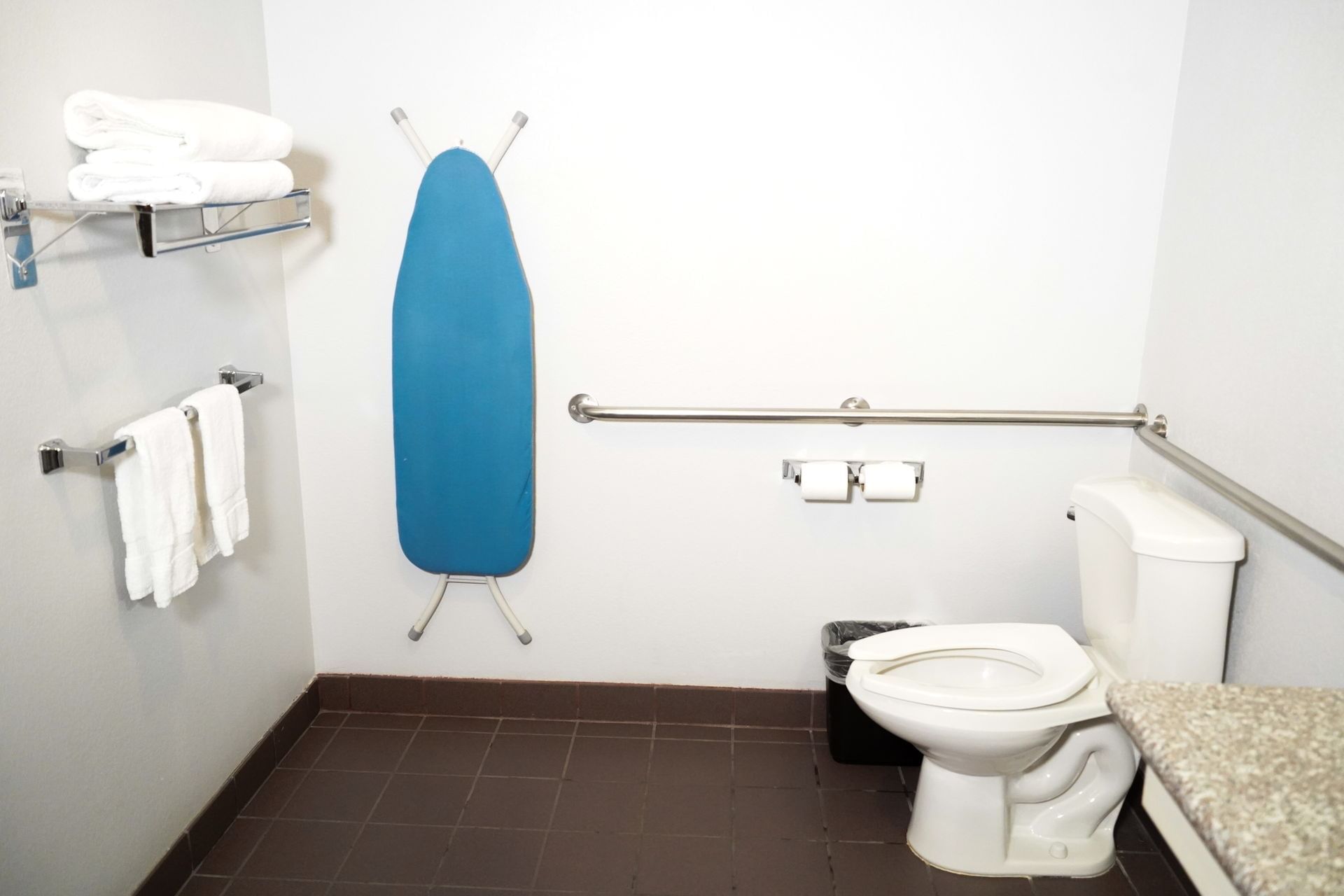 A standard hotel bathroom with a toilet, iron board, towels, and grab bars.