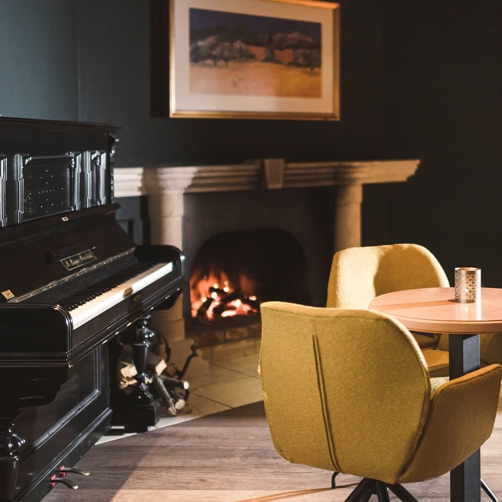 A cozy room with a fireplace, piano, framed artwork, table, and yellow chairs.