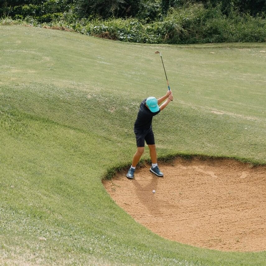 Young golfer practicing a swing from a sand bunker on a sunny golf course during PGA Junior Golf Camp.