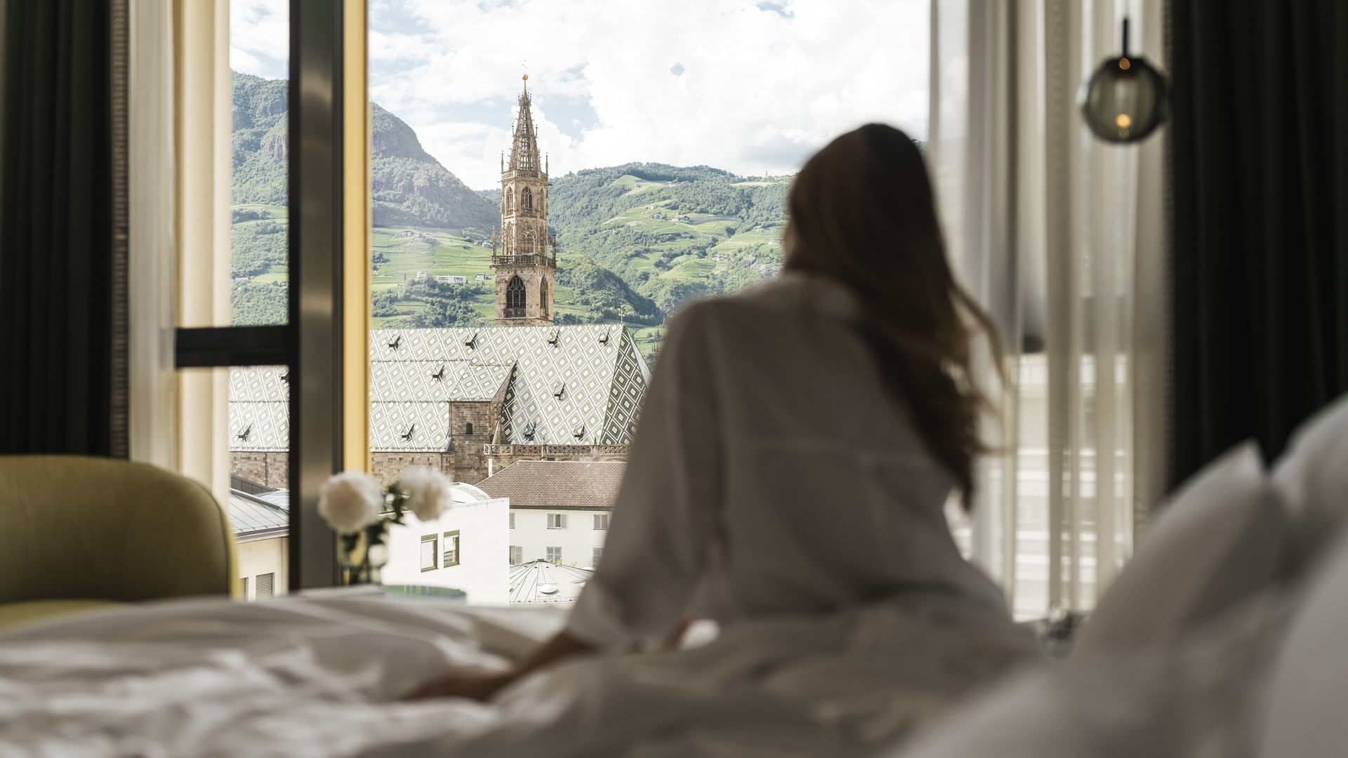 Donna seduta sul letto con vista su una chiesa e montagne.