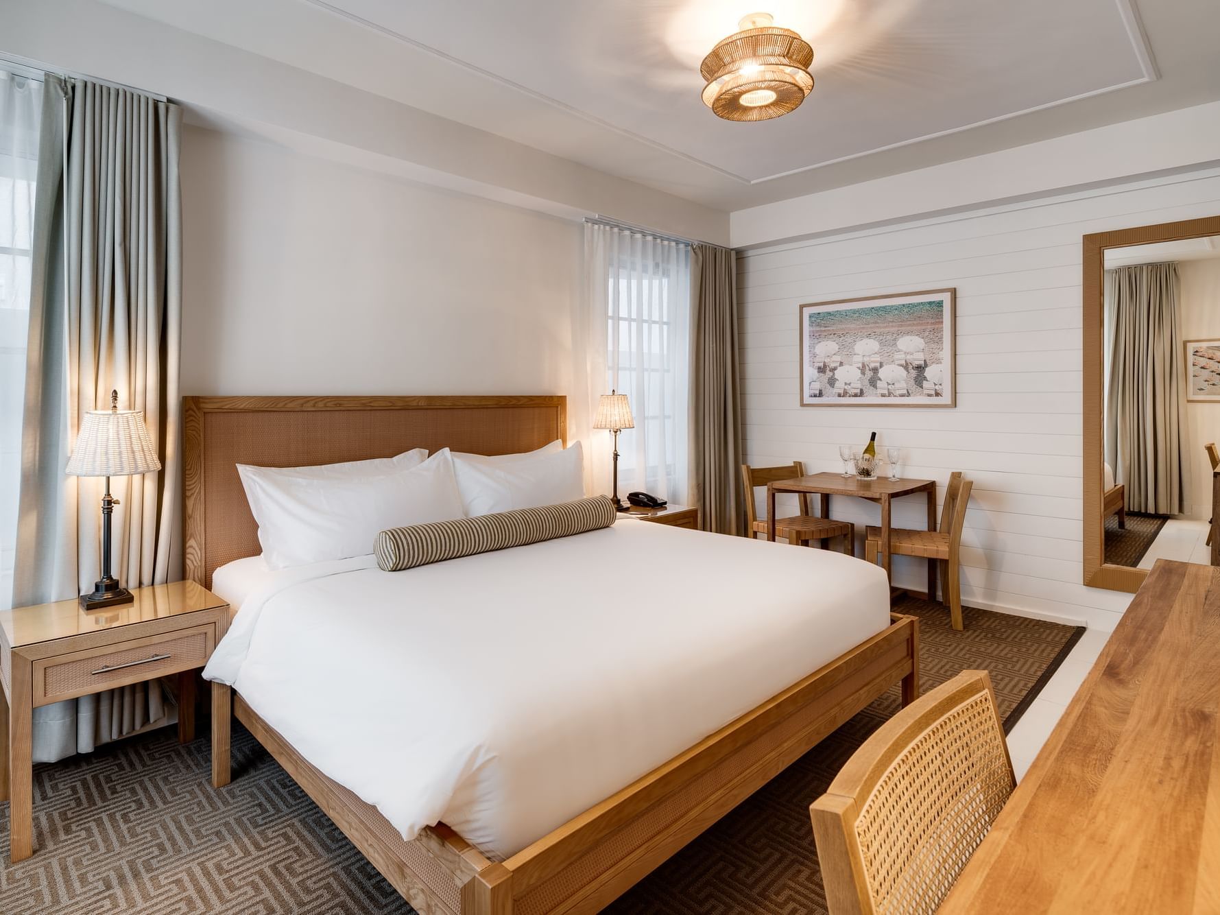 A spacious, newly renovated hotel room featuring a king bed with crisp white linens, a stylish wood desk and chair, a flat-screen TV mounted on the wall, and a small table with two chairs. The room is decorated with beach-inspired artwork and has soft lig
