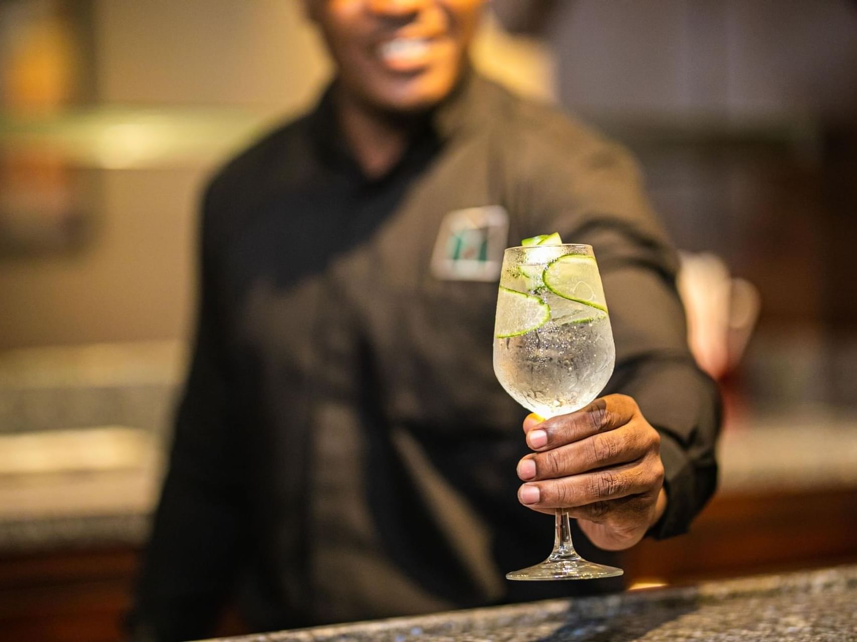Bartender in a black shirt holding out a clear cocktail glass filled with cucumber slices at Jamaica Pegasus Hotel
