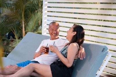 Senior couple laughing while sitting on a pool lounge chair with cocktails, next to palm trees at the Maui Coast Hotel