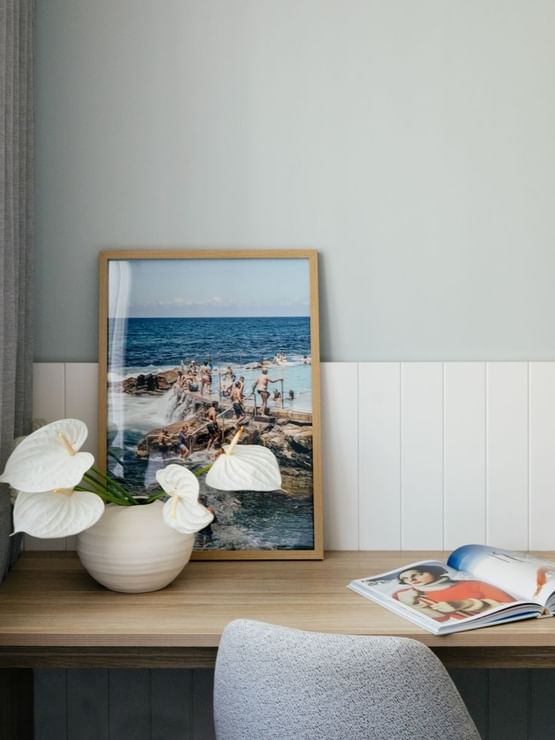 Work desk at Elysium Manly with a white vase of flowers by a coastal print, under a wall-mounted TV in Village Twin