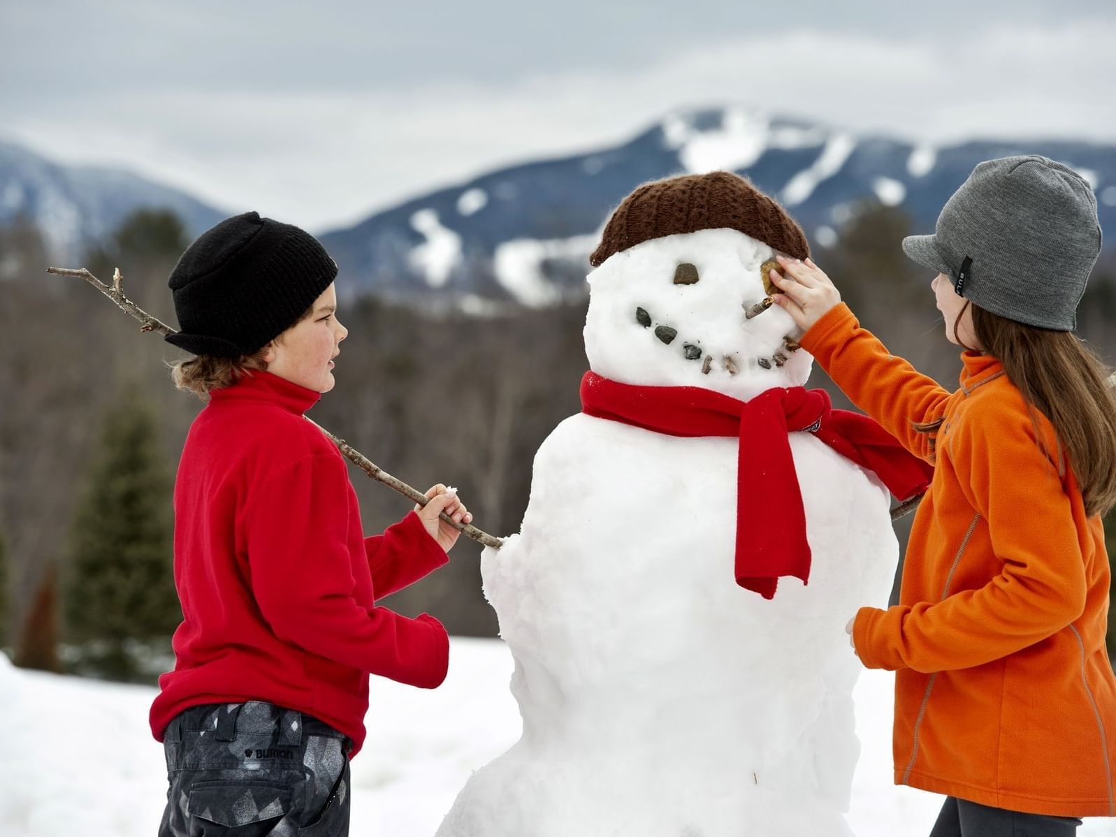 Two people building a snowman with red scarf and brown hat against snowy mountain backdrop.