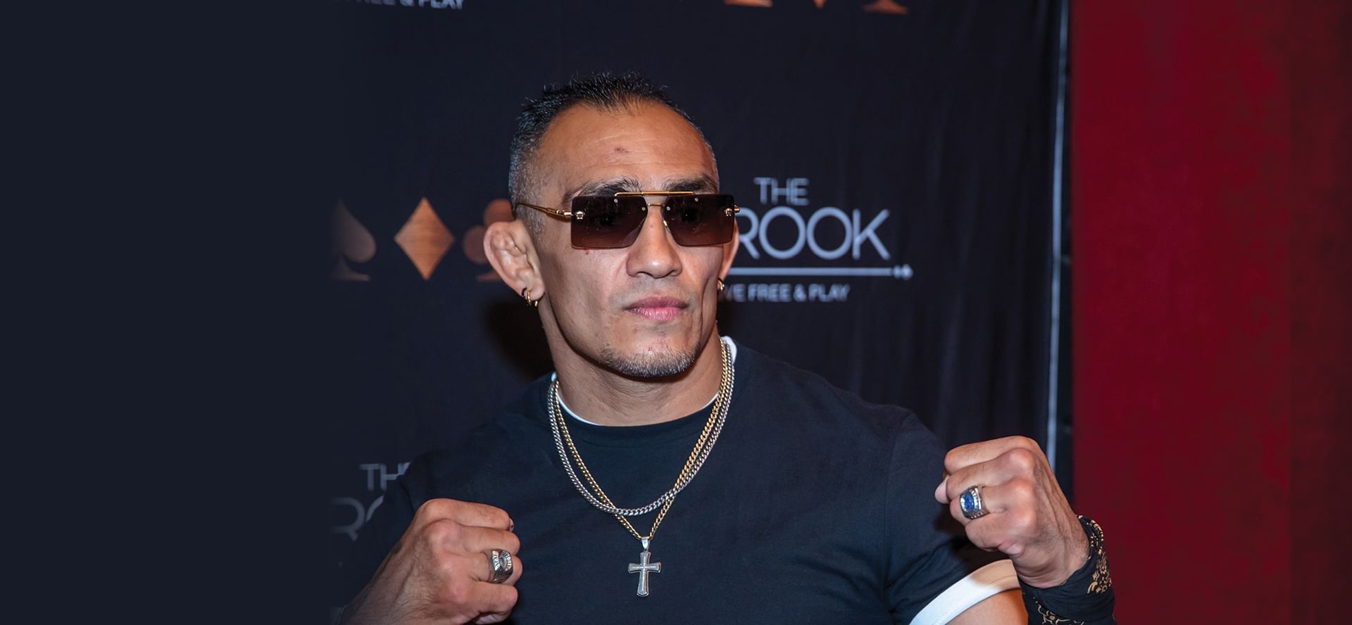 Former UFC fighter Tony Ferguson wearing sunglasses and chain necklace raising fists at event photo wall at The Brook