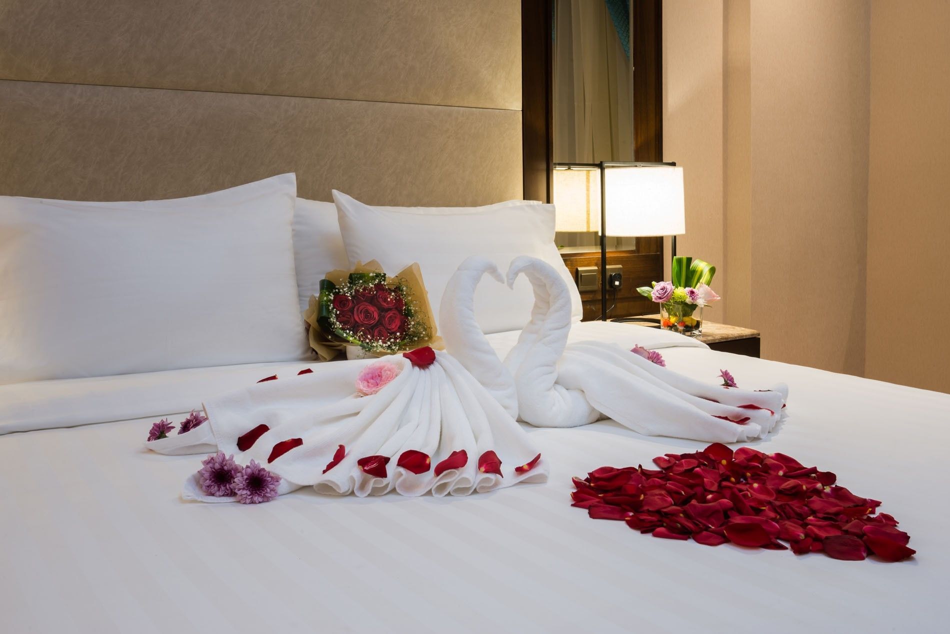 Towel swans with red rose petals on a white bed by a lamp in the Executive suite at Naviti by Warwick Dammam