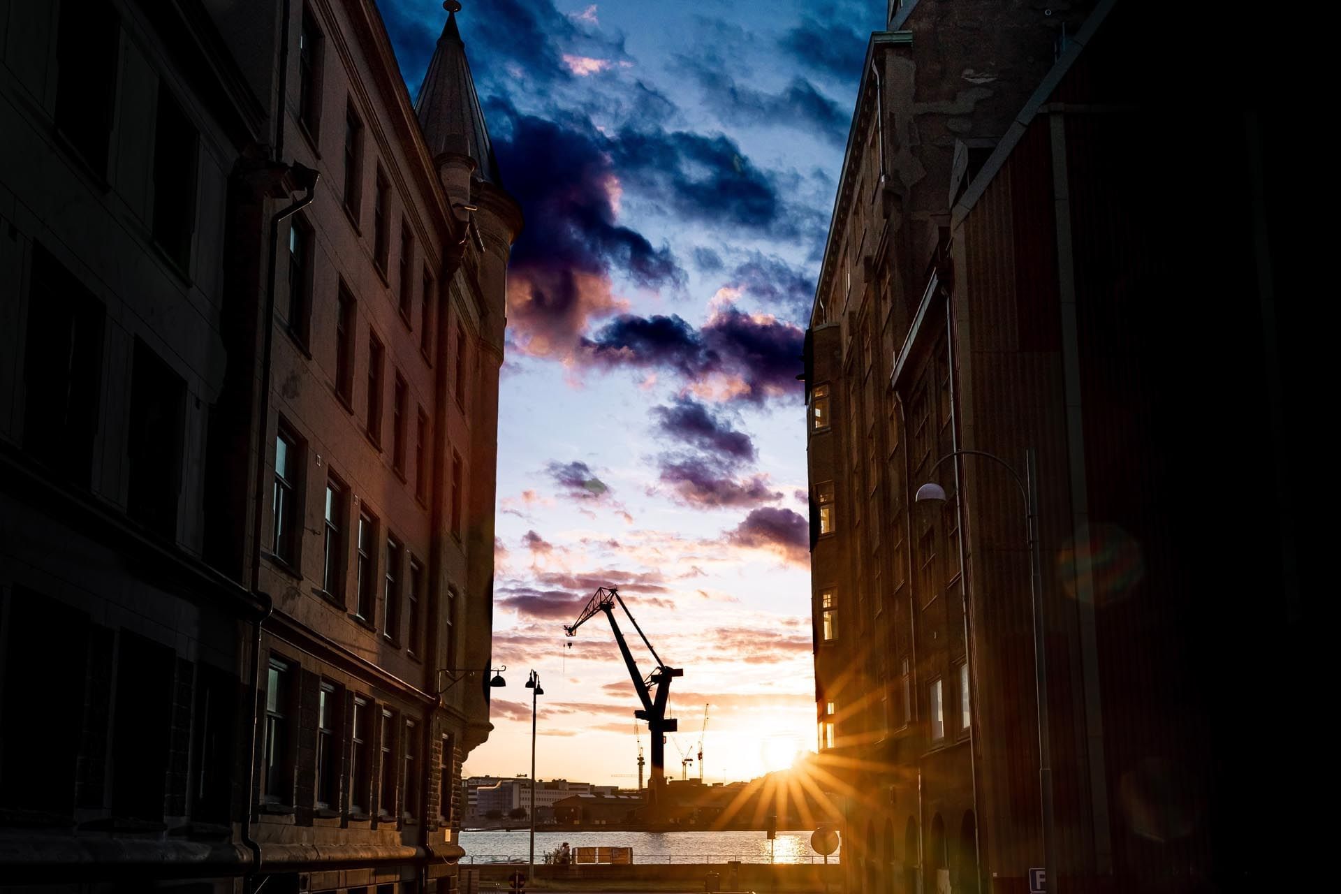 Sunset view of city buildings with crane and water in the distance, framed by tall buildings.