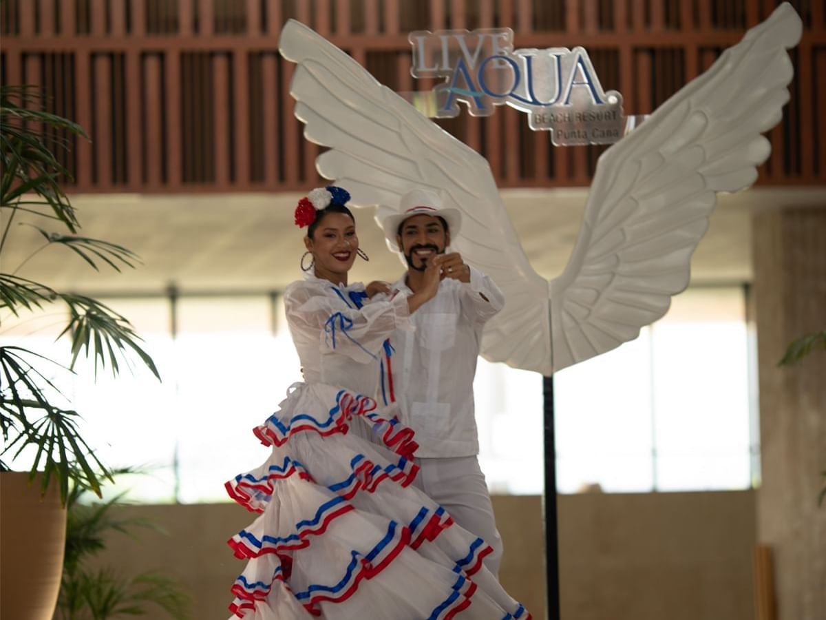 Couple dances in traditional attire near large angel wings at Live Aqua Punta Cana, showcasing colorful costumes