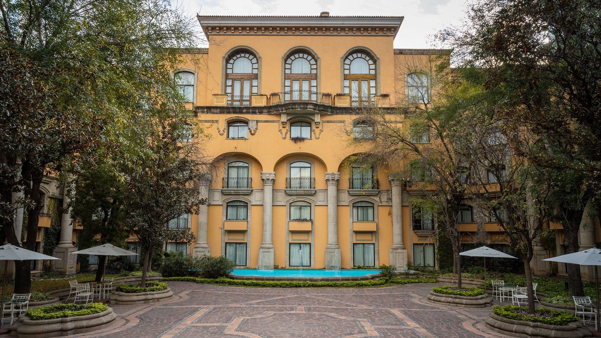 Majestic yellow facade overlooking a paved courtyard with trees and blue pool at Quinta Real Monterrey
