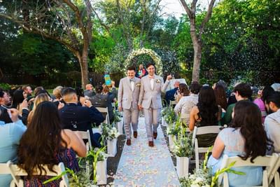 Two grooms walk hand in hand down the aisle, showered with petals during their outdoor wedding ceremony at Lake Natoma Inn