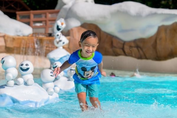 A boy in a blue bathing suit and Mickey Mouse rash guard splashes in the water with Olaf and snowgies smiling behind him.