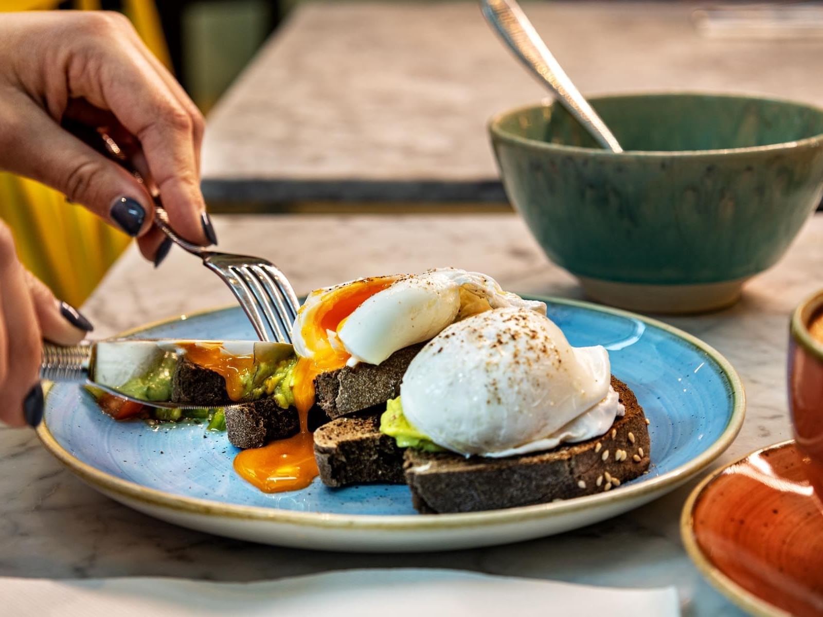 A person cutting through an avocado toast with a half-boiled egg at The Magnolia Hotel