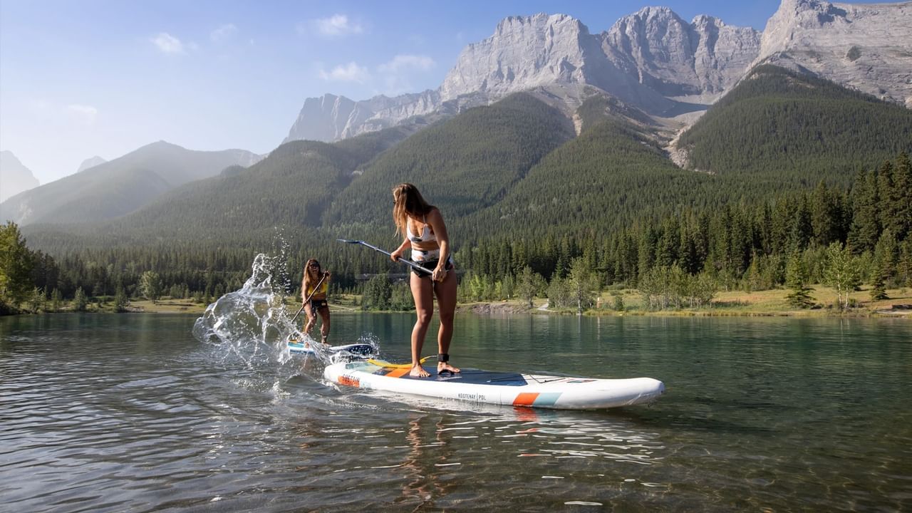 A person on a paddleboars splashes someone behind them on a lake in front of a mountain near Canmore