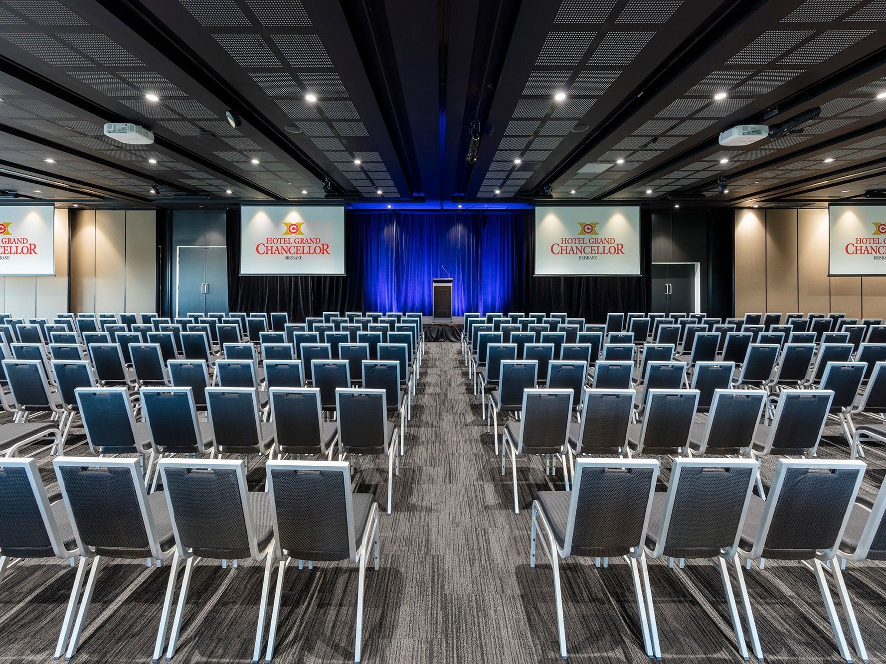 Theatre-style conference setup at Hotel Grand Chancellor Brisbane, featuring rows of seating facing a presentation stage with blue curtains and projection screens.