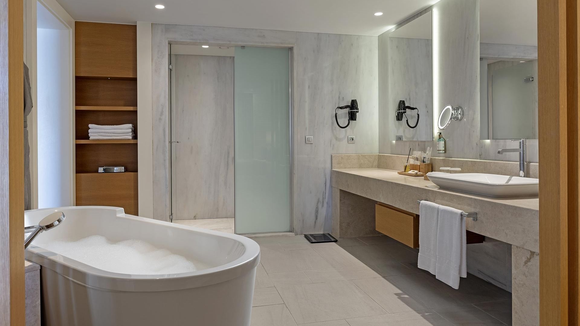 Deluxe Sea View Room's bathroom with large bathtub, double sink vanity, wooden shelves, and glass shower enclosure.