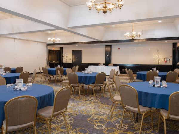 A banquet hall with round tables covered in blue cloths, chairs, and chandeliers hanging from the ceiling.