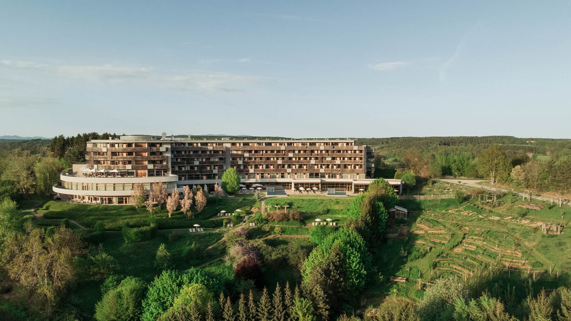 Aerial view of a large hotel building set amidst lush greenery and trees under a clear blue sky.