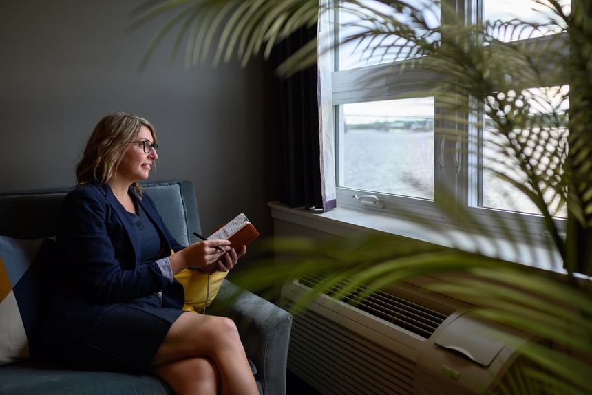 Woman sitting on a blue sofa by a window under soft drapes, with a green plant in view at Rodd Miramichi River