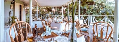 Wooden dining tables with white tablecloths and wicker chairs on a covered veranda deck at Sugar Cane Club Hotel & Spa