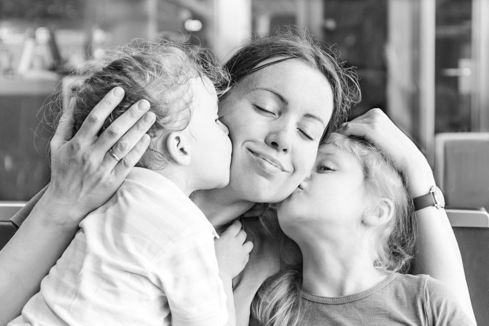 A black and white photo of a mom being kissed on either cheek by two little girls.