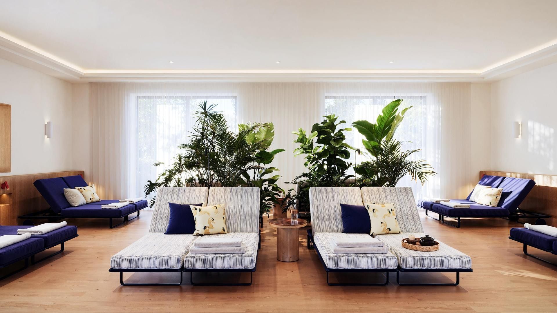 Lounge area with daybeds, pillows, and plants under large windows with white curtains.
