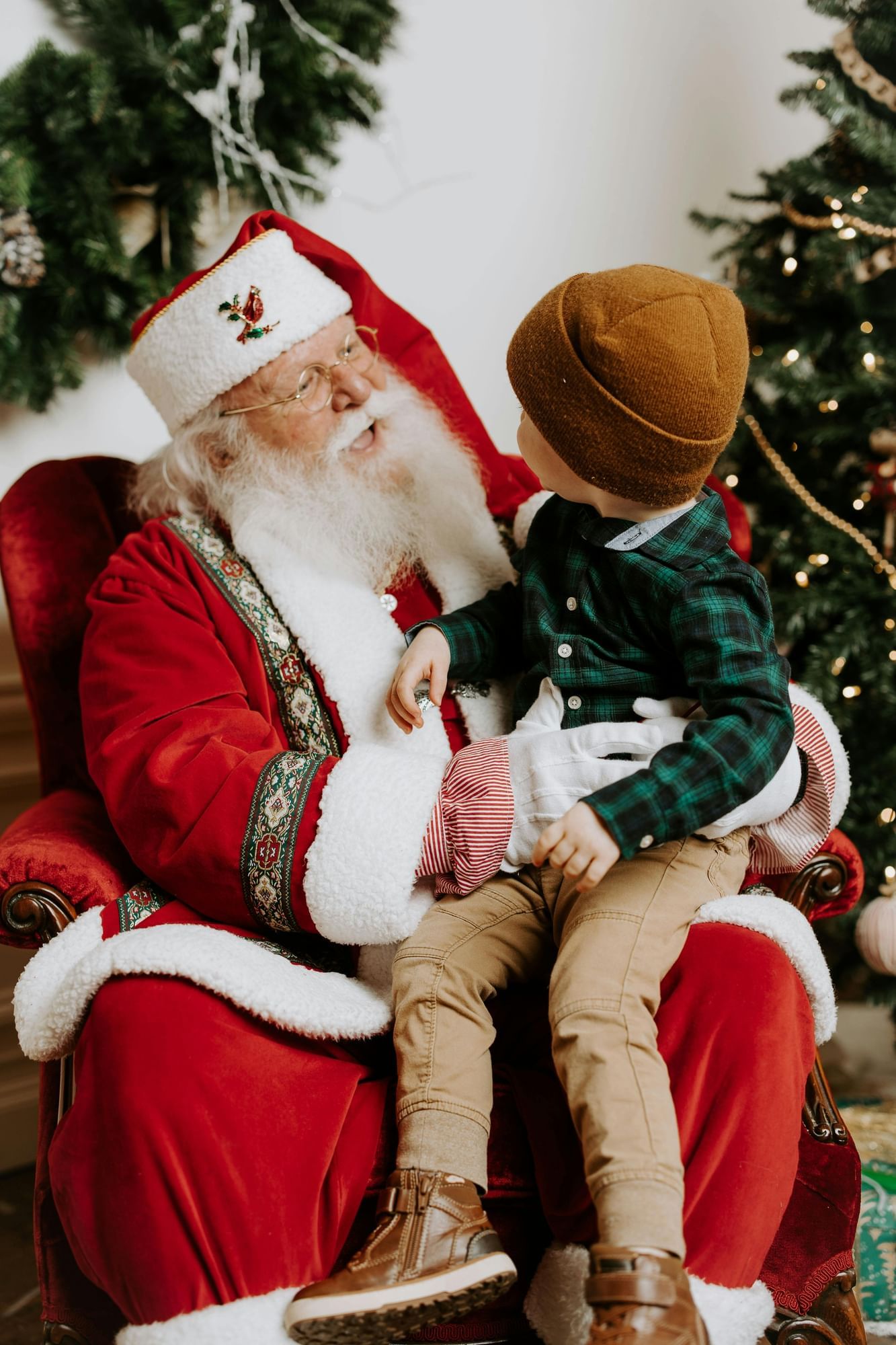 A young boy in a hat and green jacket sits on Santa's lap. 