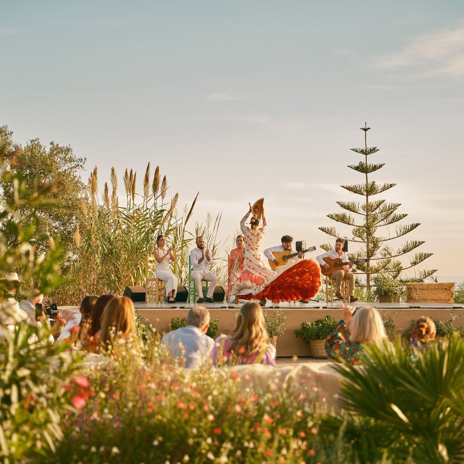Flamenco dancers and musicians performing on a stage outdoors, with audience seated among lush plants and trees.