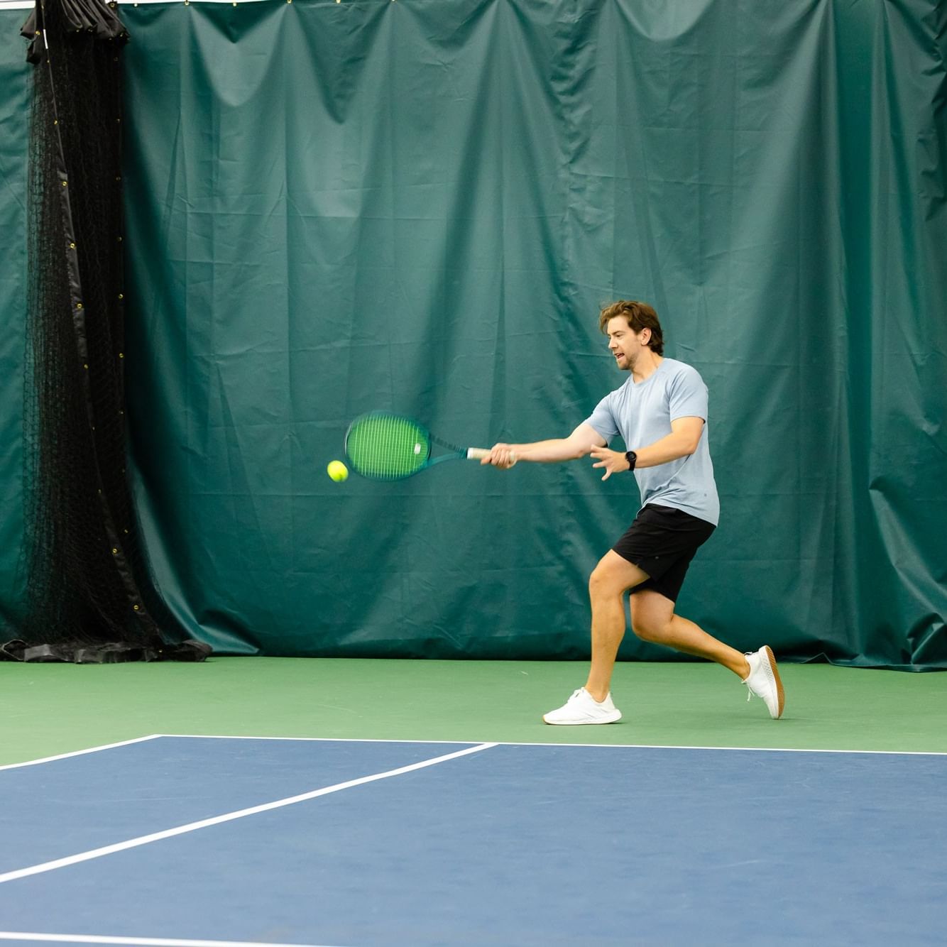 A man is playing tennis indoors on a blue court with a green net and tarp.