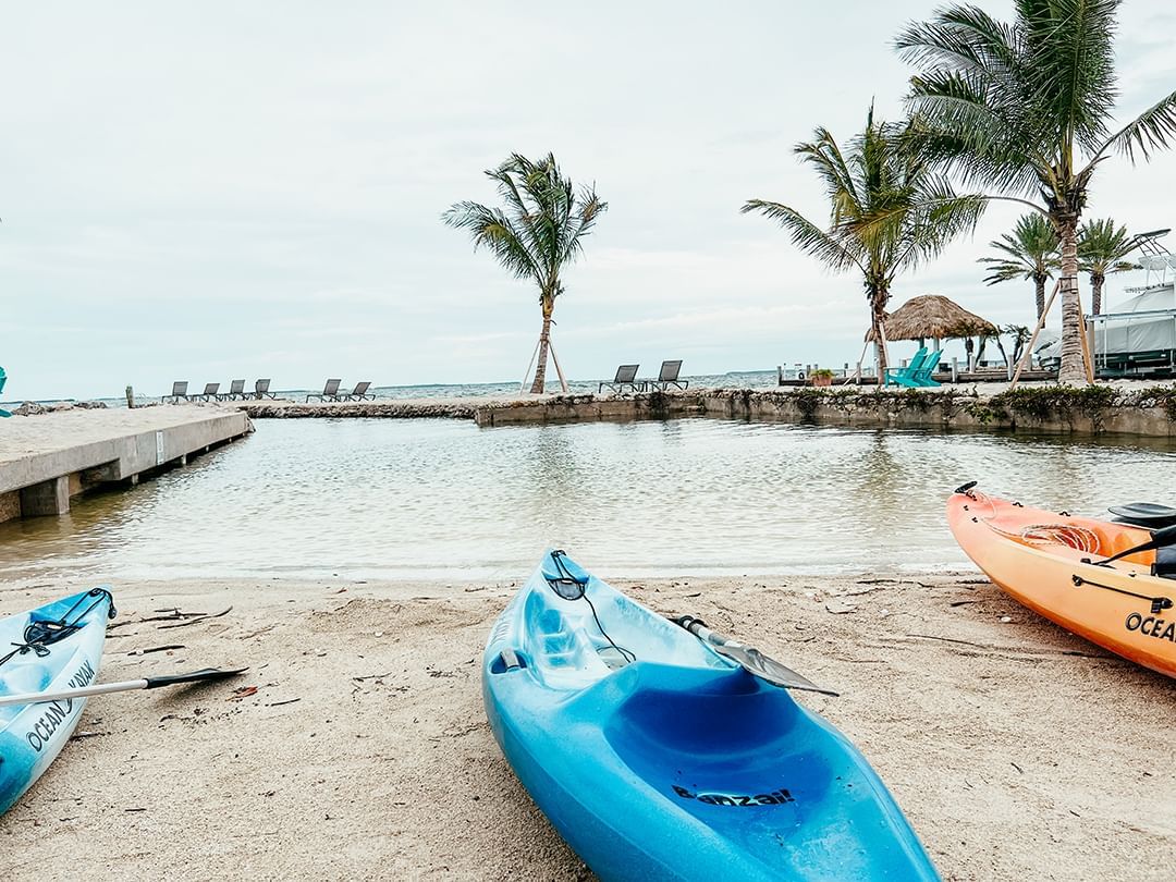 Three kayaks on sandy beach by water, with lounge chairs and palm trees in background.