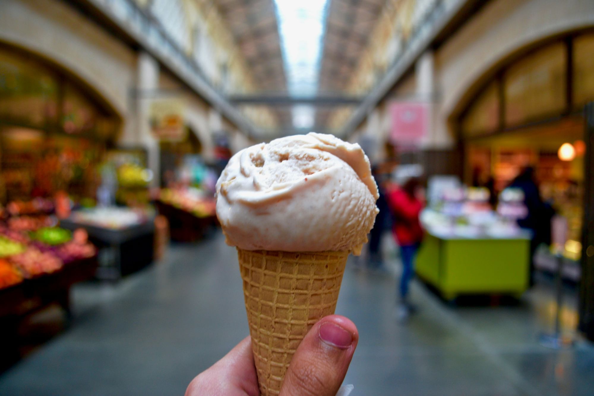 A hand holds a cone with two scoops of ice cream in front of a market background.