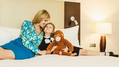 Mother and a daughter sitting on a bed with a plush monkey toy, beside a table lamp at Polonia Palace Hotel