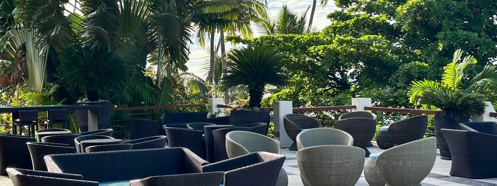Woven patio chairs by low tables under tropical palms in Sunset Terrace Bar at Warwick Fiji Resort and Spa