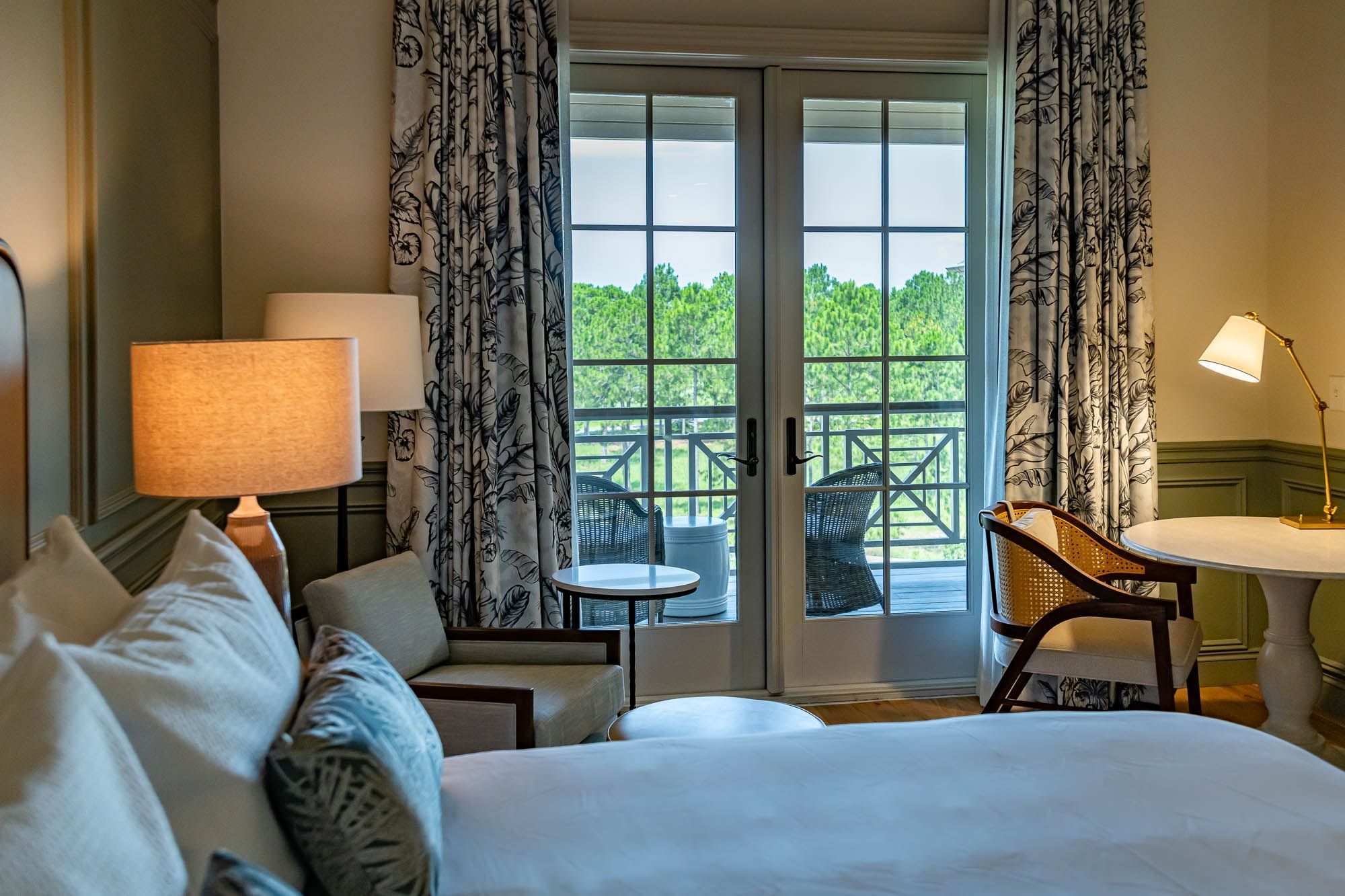 Elegant hotel room with a bed, chairs, table, and glass doors leading to a balcony with trees view.