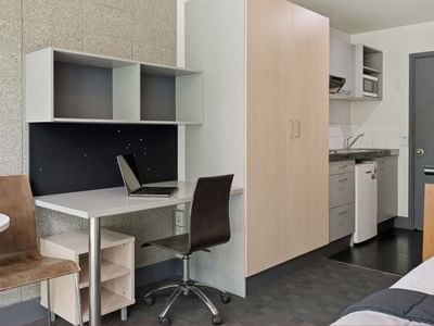 Clean, modern student living room with desk, chair, bed, and kitchen at Student Living Auckland - Beach.
