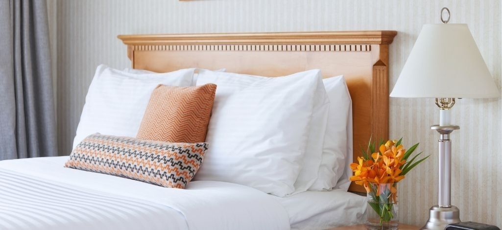 Hotel guest room with white bedding, wood headboard, patterned pillows, bedside lamp and orange flowers.