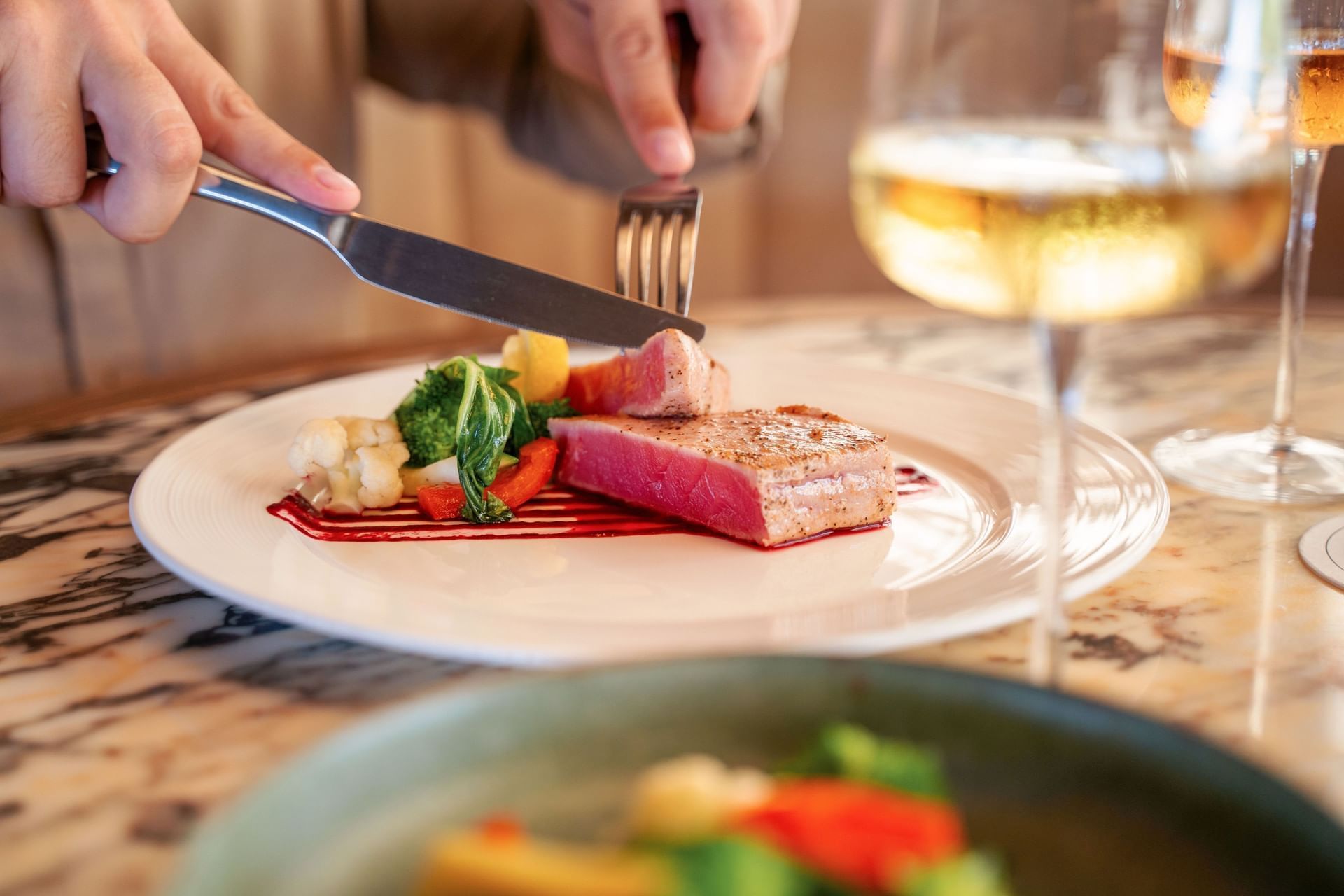 Close-up van een persoon die een tonijnsteak snijdt, gegarneerd met groenten en een saus, geserveerd bij Golden Rock Resort
