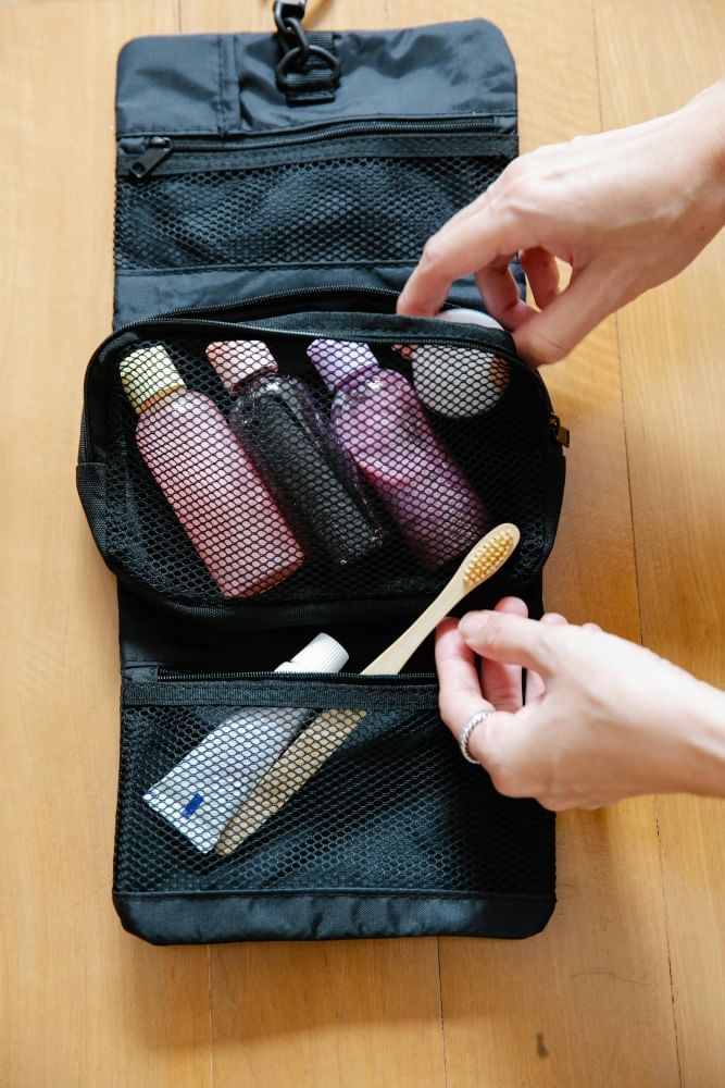 Hands put a toothbrush and travel-sized toiletries into a black toiletry bag with mesh pockets.  