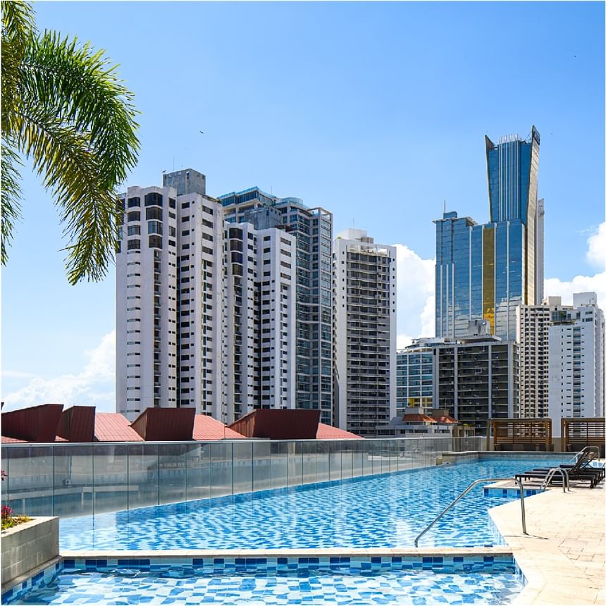 Piscina curva de azotea con barandilla de vidrio, palmera, cabañas de techo rojo y torres en Megapolis Hotel Panama