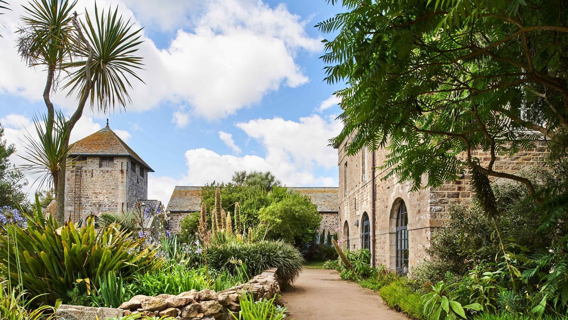Gravel walking path past tropical palms and stone archways toward historic stone castle tower at Les Maisons de Tatihou