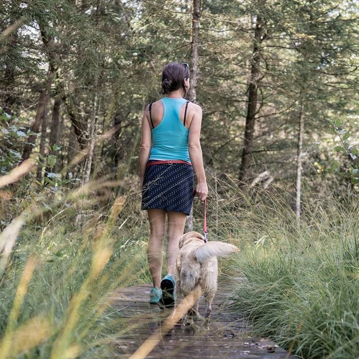 Frau in blauem Tanktop und kurzem Rock geht mit ihrem Hund im Wald spazieren.
