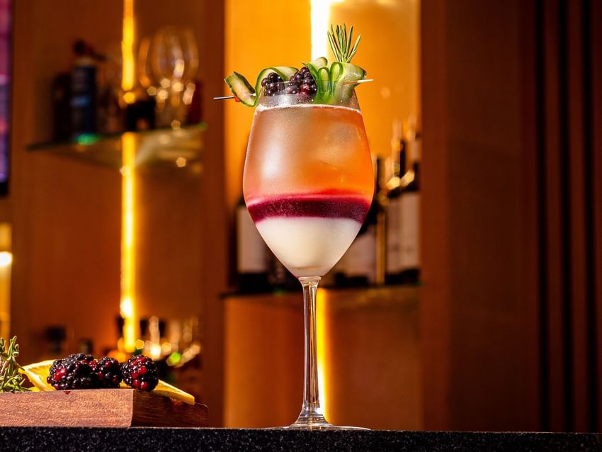 A layered fruit cocktail garnished with blackberries and cucumber in a dimly lit, elegant bar served at Camino Real