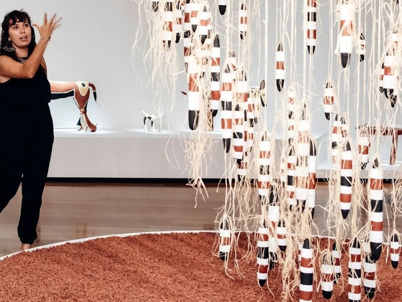 Woman gesturing near hanging textile art installation with woven elements and earth tones at QAGOMA