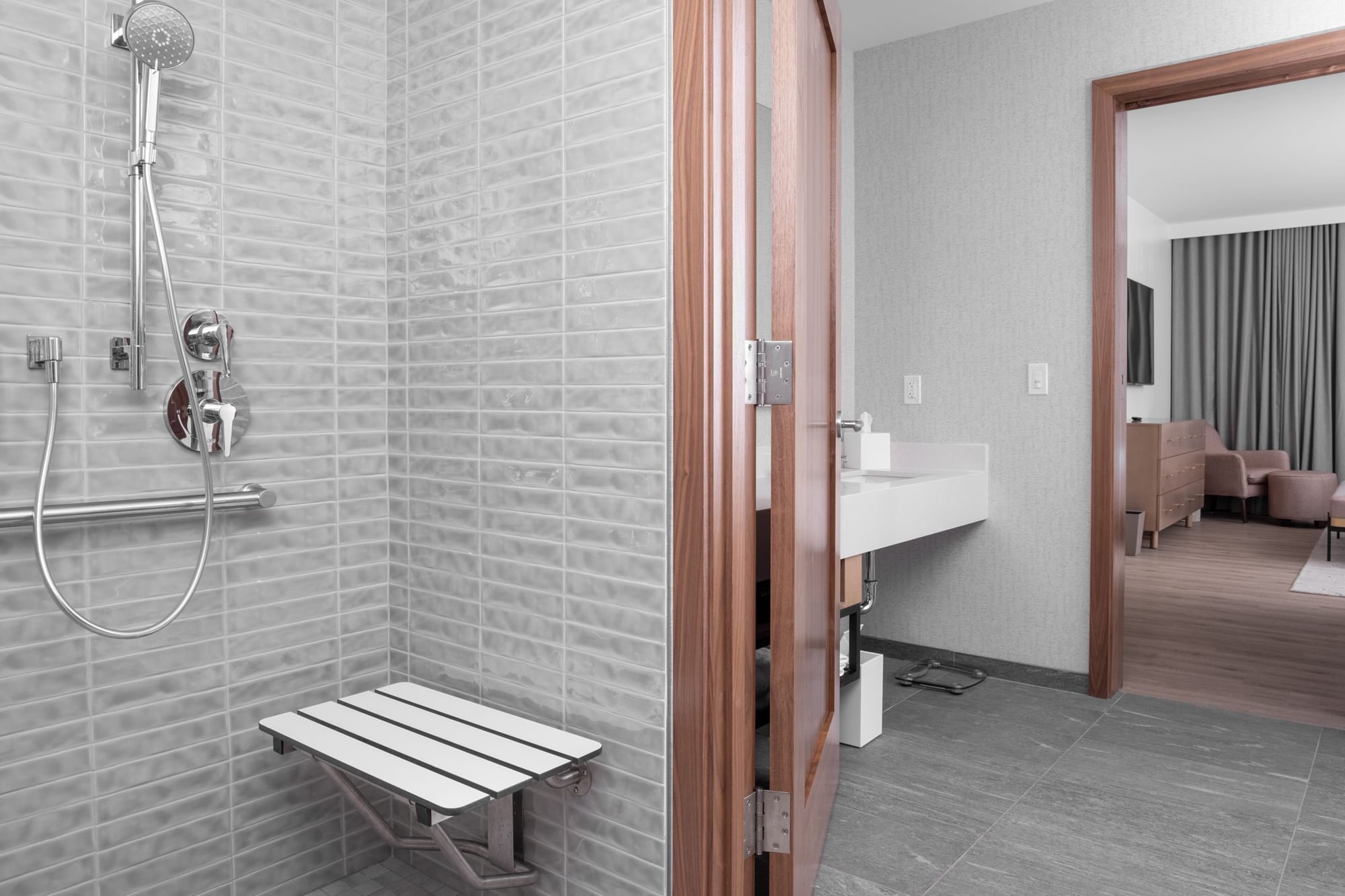A modern bathroom with a walk-in shower, bench, and rain showerhead, adjacent to a room with a bed and seating area.