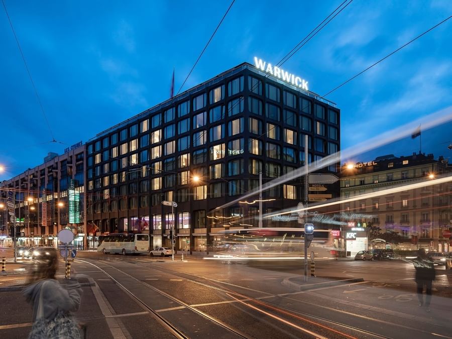 Modern glass exterior of Warwick Geneva illuminated at night on a busy city street corner during a twilight hour