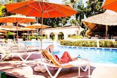 Woman relaxing on lounge chair by pool, with umbrellas and cabana, surrounded by greenery and buildings.