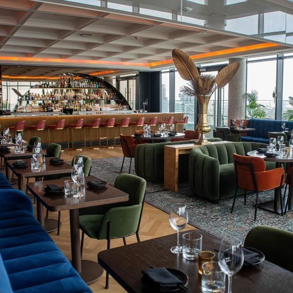 Velvet blue and green sofas by wooden tables, near a gold bar under a feather sculpture at Hotel X Toronto