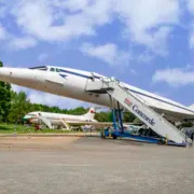 White Concorde supersonic aircraft on display with boarding stairs against a blue sky with green trees in the background.